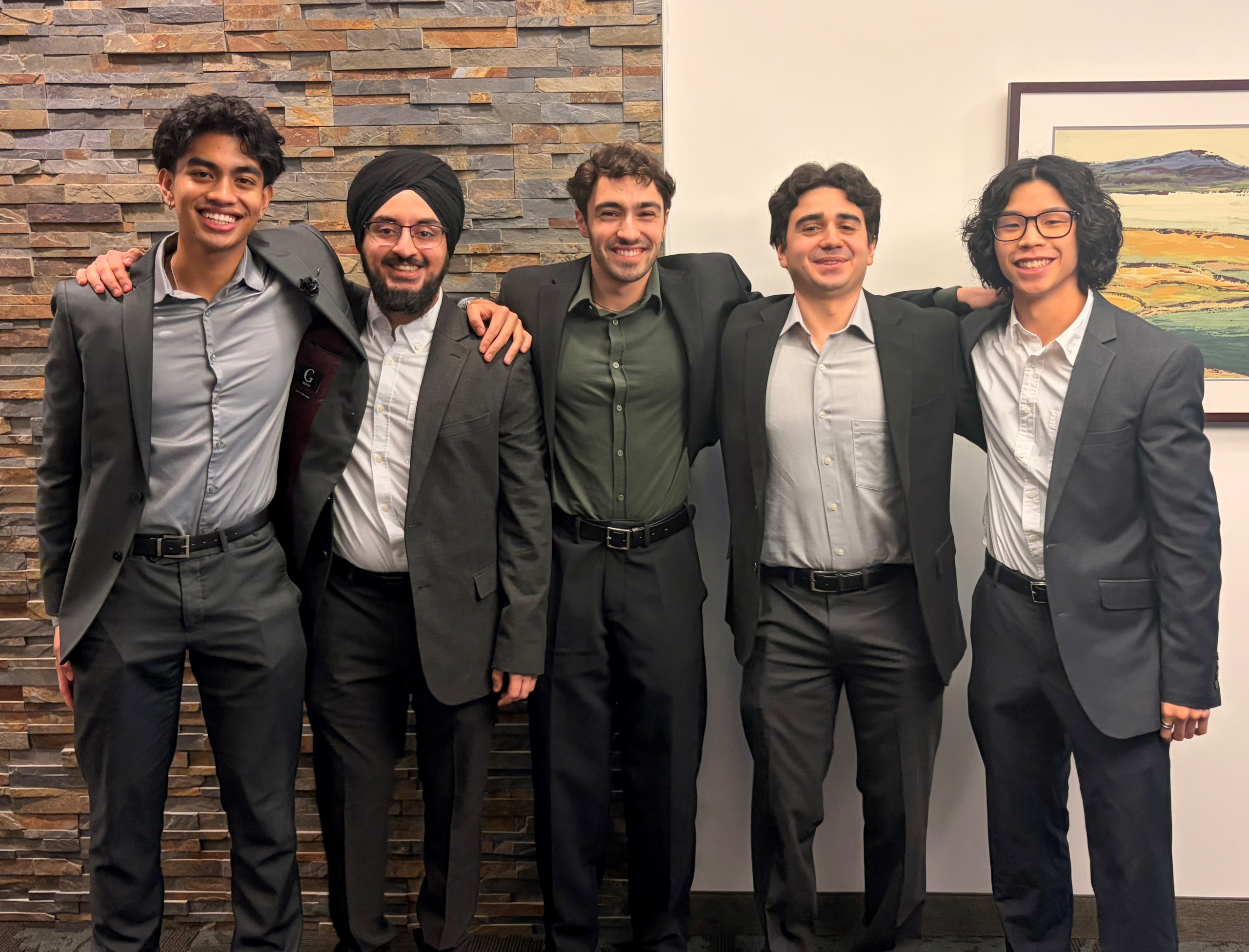 2026 Winners BioWise Solutions Five people wearing suits pose together with their arms around each other's shoulders. The group stands indoors in front of a stone accent wall and a framed landscape artwork.
